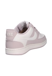 Zapatillas Nike con un diseño en blanco y rosa claro, parte superior de cuero, suela plana y un logo visible en el talón. Punta redondeada y cierre con cordones.