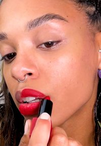 Red lipstick with a glossy finish being applied by a hand with neutral nail polish. The tube is black with a red base. Smooth skin texture.