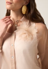 Doorzichtige beige blouse met een gelaagd stoffen bloemen detail op de borst, voorzien van witte knopen en een kraag. Gouden, gestructureerde oorbellen.