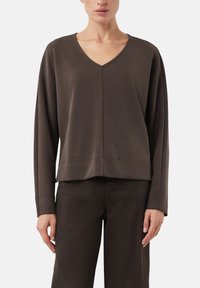 Brązowa bluzka z dekoltem w serek i długim rękawem, wykonana z miękkiego materiału, o luźnym kroju i widocznym szwie na dole. Połączona z pasującymi brązowymi spodniami.