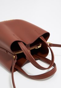 Bruine leren handtas met een gladde textuur, dubbele handvatten, ritssluiting en een ruime binnenkant. Voorzien van accenten in goudkleurig hardware.