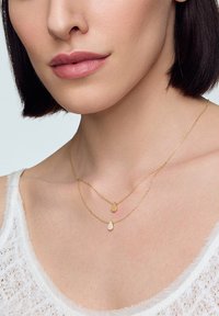Złote, warstwowe naszyjniki z zawieszką w kształcie kropli oraz mniejszą, oszlifowaną zawieszką. Łańcuszki są cienkie i delikatne na jasnym tle.