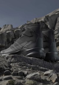 Black Adidas Terrex hiking boots placed on rugged rocky terrain under a cloudy sky, highlighting durable outdoor footwear design.