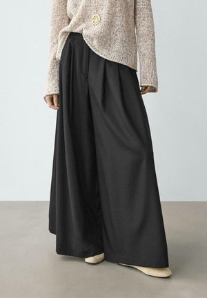 Persona che indossa un maglione beige lavorato a maglia e pantaloni neri a gamba larga, in piedi su un pavimento grigio con scarpe beige visibili.