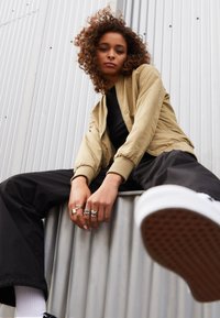 Khaki Bomberjacke mit gerippten Bündchen, schwarzem Top und lockeren schwarzen Hosen. Lockige Haare, Hände mit mehreren Ringen, im Sitzen gegen einen gewellten Hintergrund.