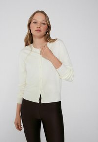 Crème-kleurige cardigan van zacht materiaal, met een ronde hals, lange mouwen en knoopsluiting aan de voorkant. Warme, gladde textuur.