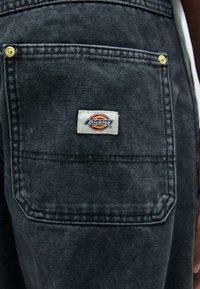 Black denim pocket with a white label featuring the "Dickies" logo in red and yellow, secured with golden metal hardware. Textured surface.