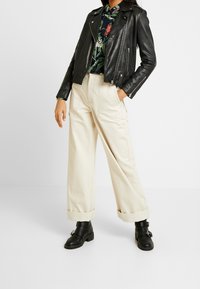 Giacca da biker in pelle nera con zip argentate, camicia con stampa floreale, pantaloni a gamba larga color crema e stivaletti neri con fibbie.