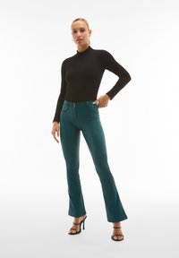 Maglione aderente nero a collo alto abbinato a pantaloni a campana turchesi con dettagli zip e doppio bottone, completato da tacchi neri con cinturino.