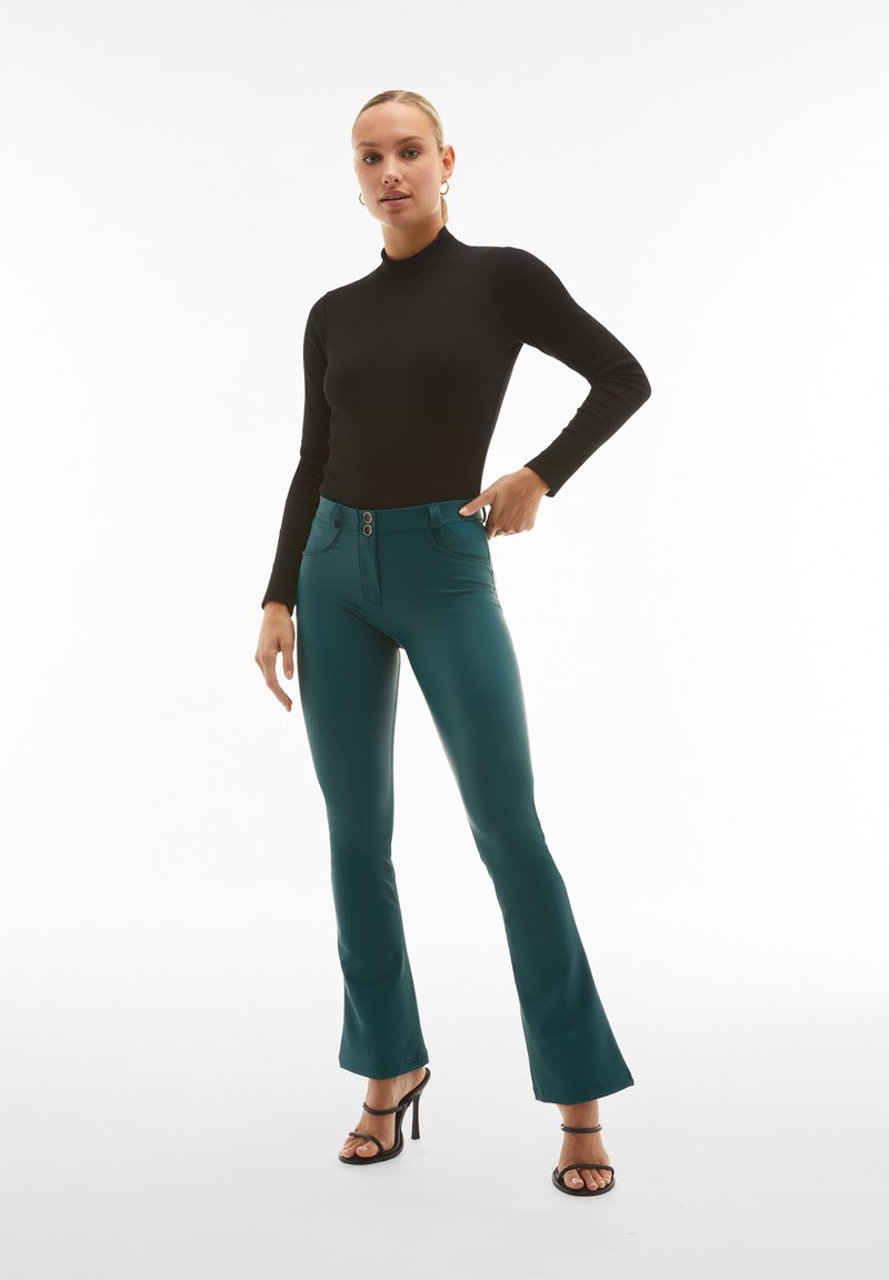 Maglione aderente nero a collo alto abbinato a pantaloni a campana turchesi con dettagli zip e doppio bottone, completato da tacchi neri con cinturino.