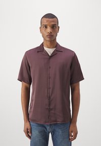 Chemise à manches courtes de couleur bordeaux en tissu lisse, avec un col en V, des boutons sur le devant et un ourlet droit. Associée à un jean bleu.