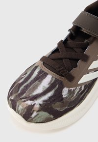 Pantof sport cu model camuflaj, având o parte superioară din țesătură texturată în nuanțe pământii, șireturi maro și o talpă albă cu accente minimaliste.