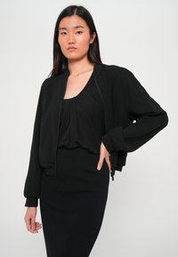 Mustat pommittajatakki, jossa on sileä pinta, joustavat hihansuut ja vetoketjukiinni. Päällä musta paita ja slim fit -musta hame.