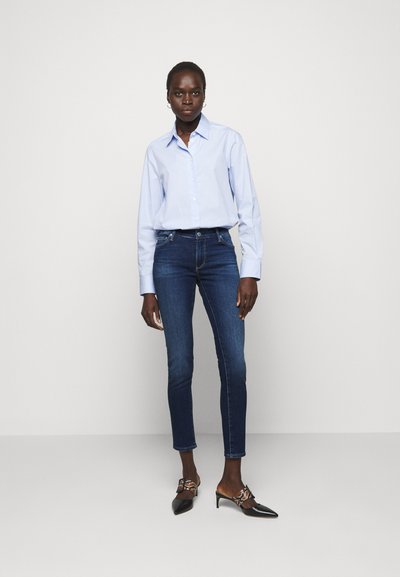 Camisa azul clara de botão, jeans skinny azul escuro e sabrinas pretas de bico fino com fivela decorativa. Tecido suave de mistura de algodão.