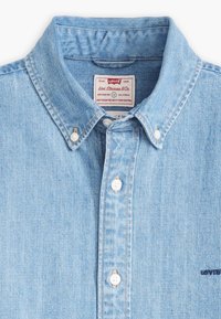 Light blue denim button-down shirt with a pointed collar, white buttons, and a small dark blue logo on the pocket. Tag visible inside collar.