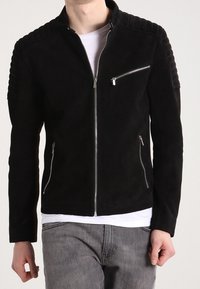 Giacca in suede nera caratterizzata da un design aderente, profondi accenti sulle spalle e cerniere argentate sul petto e nelle tasche laterali. Collo semplice.