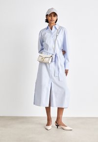 Jasnoniebieska, długa koszula z długim rękawem w formie sukienki z wiązanym pasem, z bocznym rozcięciem, zestawiona z białą torebką na ramię i kremowymi szpilkami.