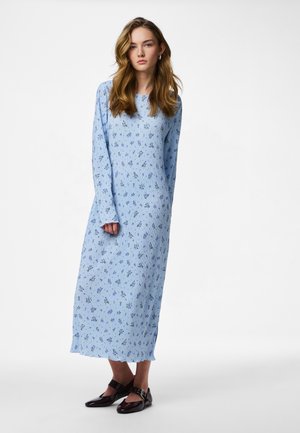 Γυναίκα με μακριά κυματιστά μαλλιά που φοράει γαλάζιο μακρύ φόρεμα με μακριά μανίκια και μικρά μωβ λουλουδάκια, καθώς και μαύρα παπούτσια τύπου Mary Jane.