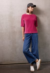 Top corto a righe rosa con maniche corte, jeans a gamba larga blu e scarpe da ginnastica bianche con suola beige. Modella che indossa un cappellino da baseball scuro, in piedi contro un muro di cemento.