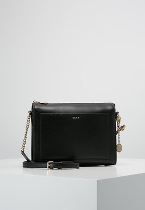 Bolso bandolera de cuero negro DKNY con cadena dorada, cierre de cremallera y charm redondo dorado sobre una superficie blanca.