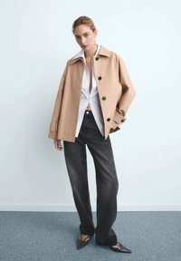 Béžová oversized bunda s černými knoflíky, bílá košile pod ní a tmavé džíny. Špičaté hnědé boty, stojící na šedém koberci.