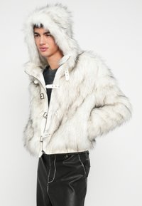 Pessoa jovem vestindo casaco branco com capuz de pelo sintético e calças pretas de couro, em pé com as mãos nos bolsos, voltada ligeiramente para a esquerda.