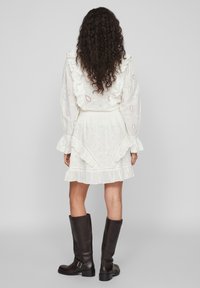 Žena s kudrnatými vlasy, oblečená v bílých šatech s volány a výšivkou, v černých kožených kozačkách až po kolena, stojící zády.