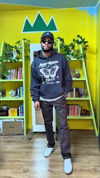 Felpa nera con grafica di una farfalla, camicia beige, pantaloni marroni e scarpe da ginnastica bianche. Lo sfondo presenta scaffali verdi e pareti gialle.