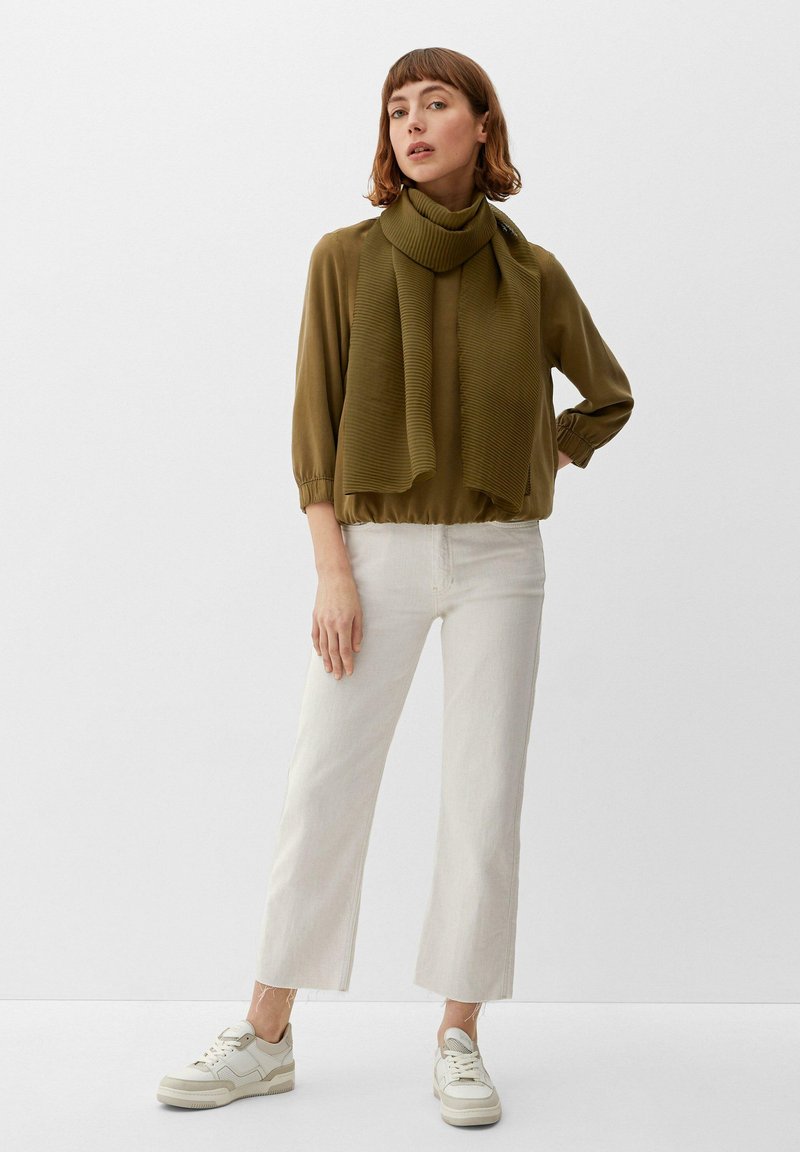 Blouse vert olive avec une écharpe côtelée, manches trois-quarts et silhouette cropped, associée à un pantalon large blanc et des baskets crème.