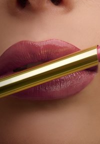Labbra glossy color malva che tengono un tubetto di rossetto oro con una tonalità malva abbinata vicino al labbro inferiore.