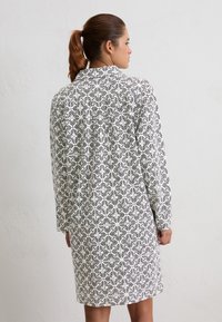 Mujer con una coleta castaña, vistiendo un vestido blanco de manga larga con un patrón cuadrado de flores negras, de pie contra una pared clara y lisa.