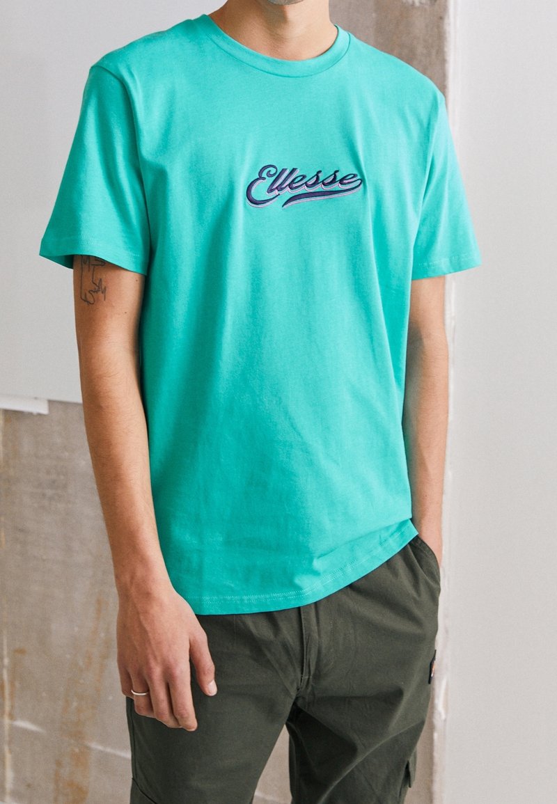 T-shirt turquoise à manches courtes en coton avec le logo "Ellesse" brodé en violet sur la poitrine ; encolure ronde, coupe décontractée.