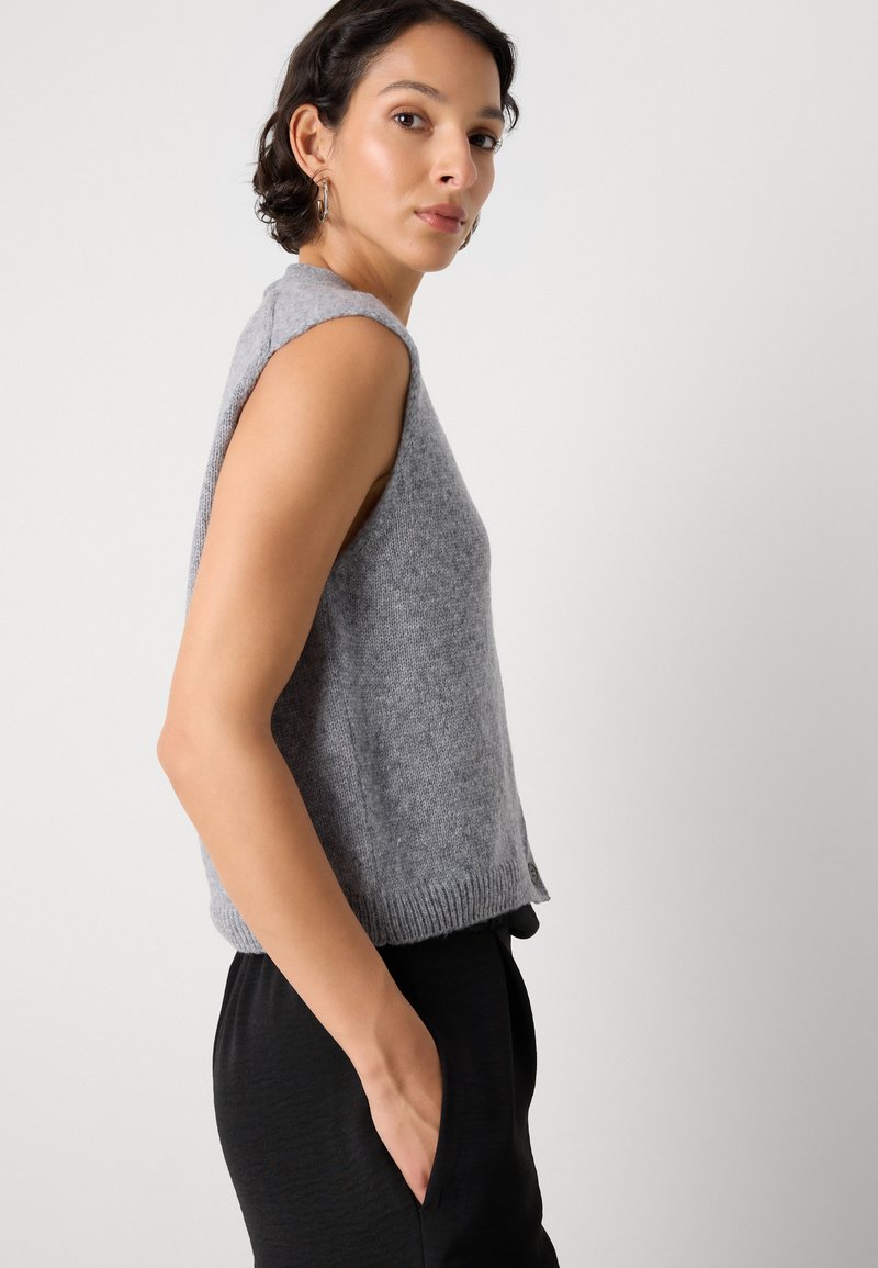 Hellgrauer ärmelloser Pullover, gestrickte Textur, runder Hals, gerippter Saum. Getragen mit schwarzen Hosen, zeigt das Seitenprofil und eine lockere Passform.