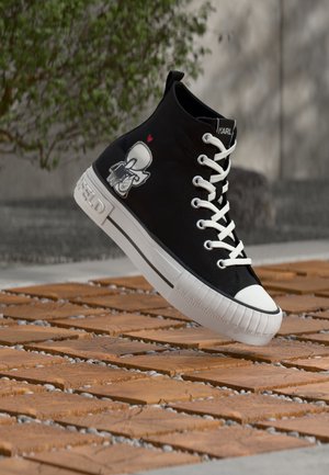 Black high-top sneaker with white laces and sole, cartoon character illustration on side, floating above wooden-patterned ground outdoors.