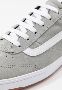 Baskets en daim gris avec des lacets blancs et une bande latérale en cuir blanc. Présente une surface texturée et une semelle en caoutchouc, avec des détails de couture visibles.