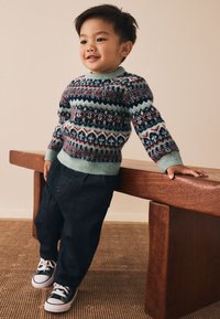 Flerfärgad tröja med mönster och turkosa detaljer, mörkblå jeans och marinblå sneakers. Står vid en bänk i trä.