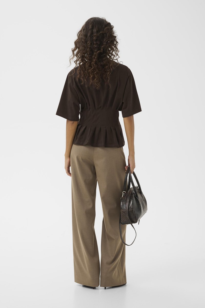 Haut peplum marron à manches courtes, associé à un pantalon beige taille haute. Tenant un sac à main noir texturé. Cheveux bouclés visibles.