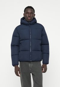 Tamsiai mėlynas pūkinis švarkas su gobtuvu, užsegamas zipu, quilted dizaino ir elastiniais rankogaliais, derinamas su pilkais džinsais.