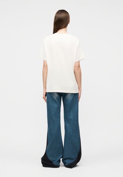 Άτομο με μακριά καστανά μαλλιά φοράει απλό λευκό κοντομάνικο πουκάμισο και φαρδιά μπλε τζιν με μαύρα πάνελ, στέκεται μπροστά σε απλό φόντο.