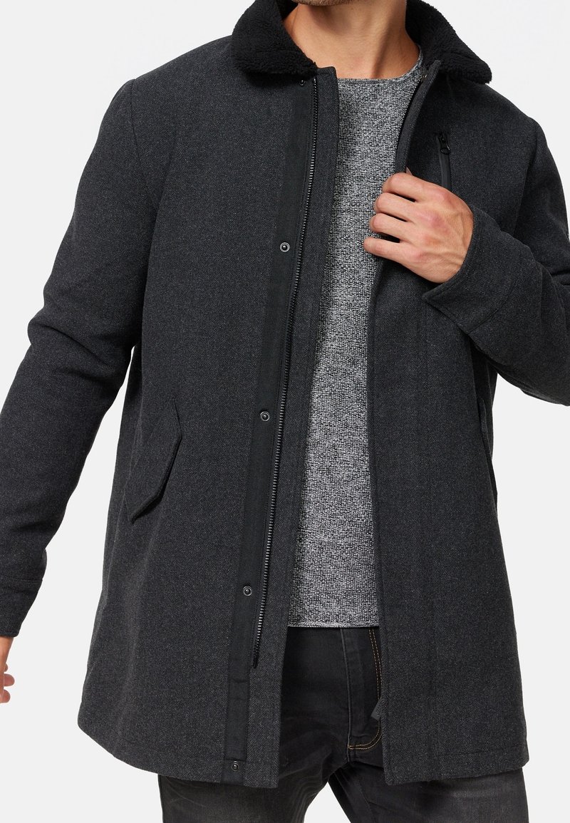 Manteau gris foncé en tissu texturé, avec un col doublé de fourrure noire, une fermeture éclair avant et des poches latérales. Porté par-dessus un pull gris clair.