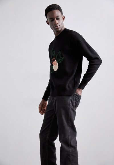 Black sweater with floral embroidery in reds and greens. Worn with gray jeans. Sweater features a round neckline and ribbed cuffs.