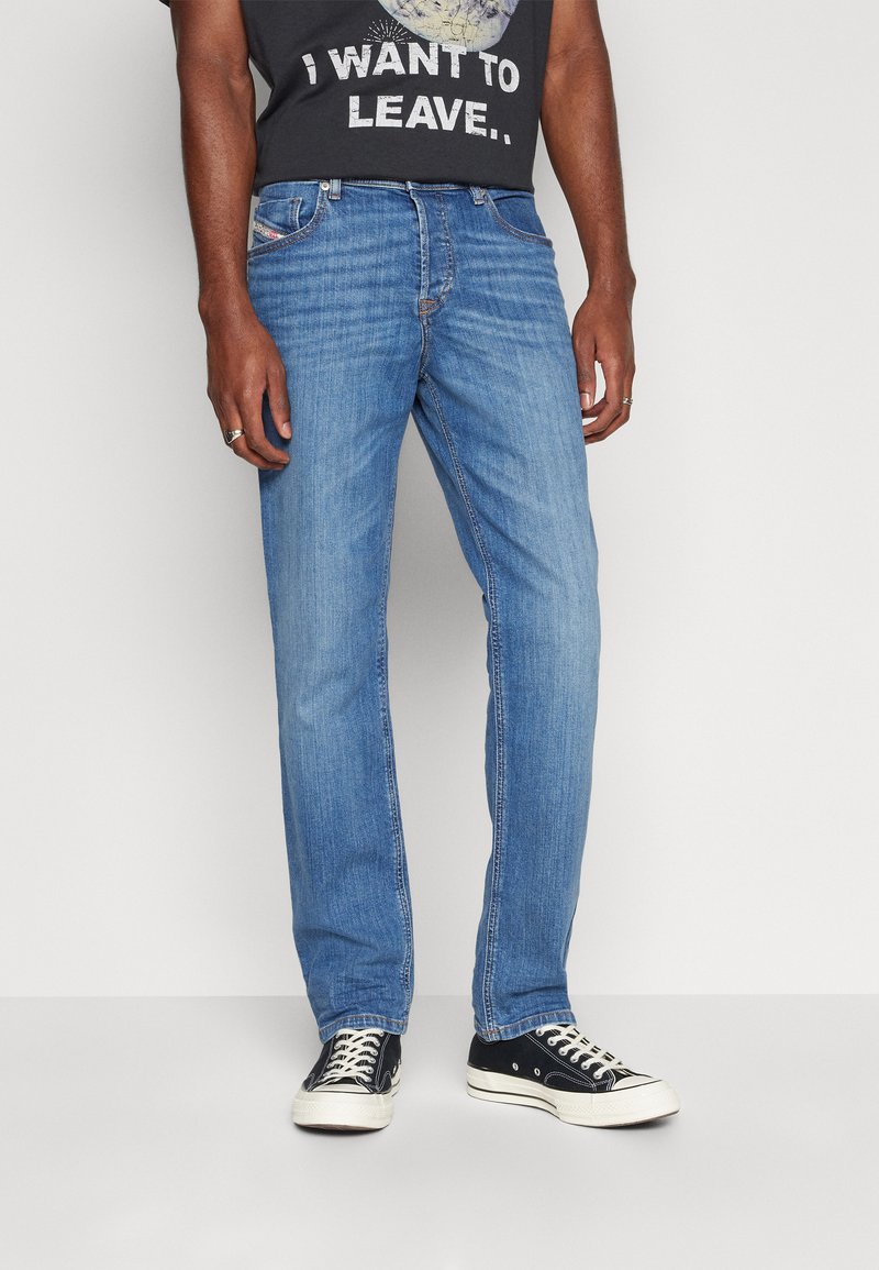 Helle blaue Jeans mit geradem Schnitt, ausgestattet mit fünf Taschen, dezenten Stickdetails und einem leichten Verblassen. Getragen mit schwarzen Sneakern.