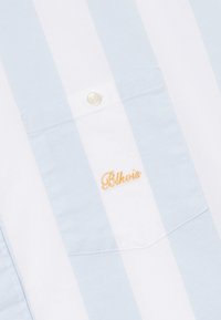 Chemise rayée verticale bleu clair et blanc avec un bouton et le texte brodé jaune "Blkvis" sur la poche poitrine.