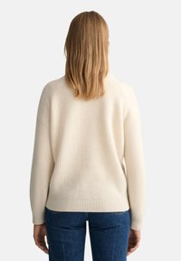 Suéter de punto color crema con textura acanalada, ajuste relajado y hombros caídos, combinado con jeans azules. Vista trasera que muestra un diseño sin costuras.