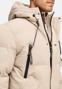 Chaqueta acolchada de color beige con capucha, que presenta herrajes negros, cordones ajustables y un bolsillo con cremallera. Detalle de tela texturizada visible.