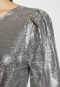 Silver paljettopp med texturerad yta, puffärmar och V-ringning. Glittrande paljetter ger en reflekterande effekt.