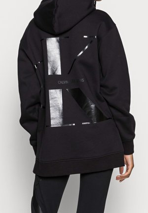 Osoba ubrana w czarną, oversize'ową bluzę z błyszczącym, czarnym geometrycznym wzorem i napisem „Calvin Klein Jeans” na plecach, w połączeniu z czarnymi legginsami.