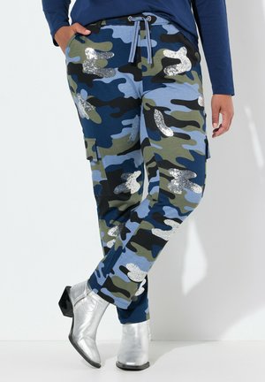 Camo cargo pants with green, blue, and black pattern. Features silver sequined accents and a drawstring waist. Worn with silver heeled boots.