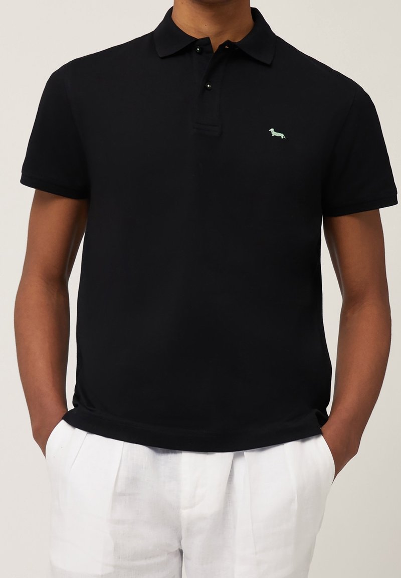 Homme portant un polo noir à manches courtes avec un petit logo chien vert clair sur la poitrine et un pantalon blanc, mains dans les poches.