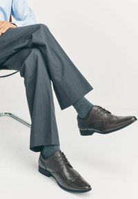 Chaussures habillées en cuir marron avec un design brogue, présentant une finition lisse, un bout arrondi et des lacets. Portées avec des chaussettes grises et un pantalon gris.